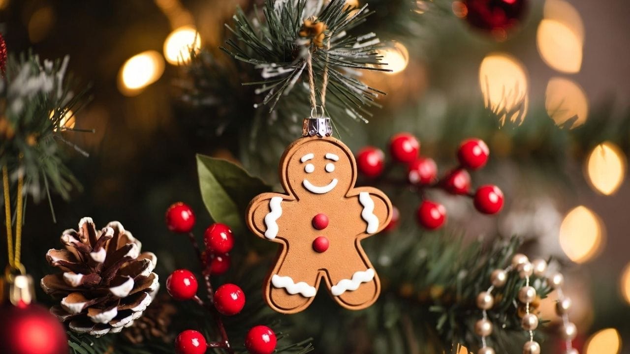 Gingerbread man ornament hanging on a Christmas tree surrounded by pine needles, red berries, a frosted pinecone, and warm golden holiday lights in the background.